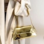 Faites briller votre style avec le sac à main Molly en vernis doré, une pièce luxueuse portée avec délicatesse au bras, ajoutant une touche glamour à votre look.