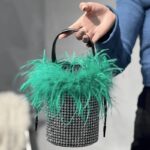 Élégance féminine incarnée : le sac à main Kendal, orné de strass et de plumes vertes, tenu avec grâce par une femme.