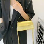 Sac à main Nawel doré en similicuir brillant, porté sous le bras par une femme.
